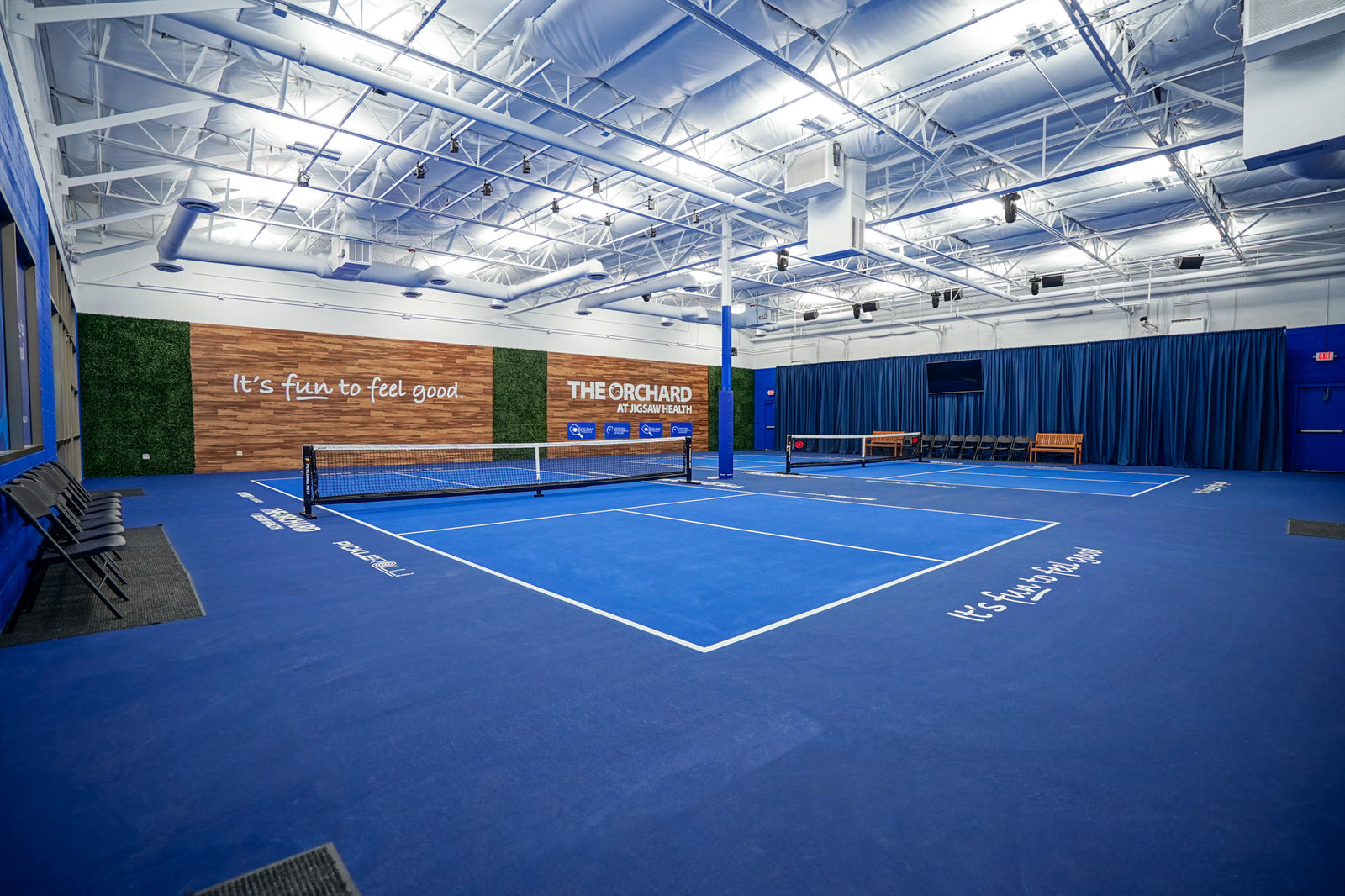 Indoor pickleball courts at The Orchard by Jigsaw Health with blue flooring and branded wall reading “It’s fun to feel good.”
