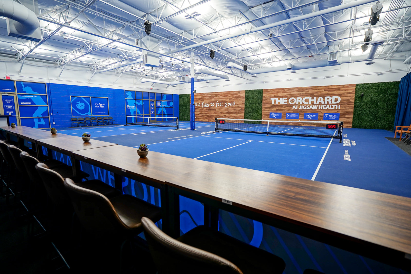 View of The Orchard at Jigsaw Health pickleball courts with courtside seating, high-top chairs, and branded walls.