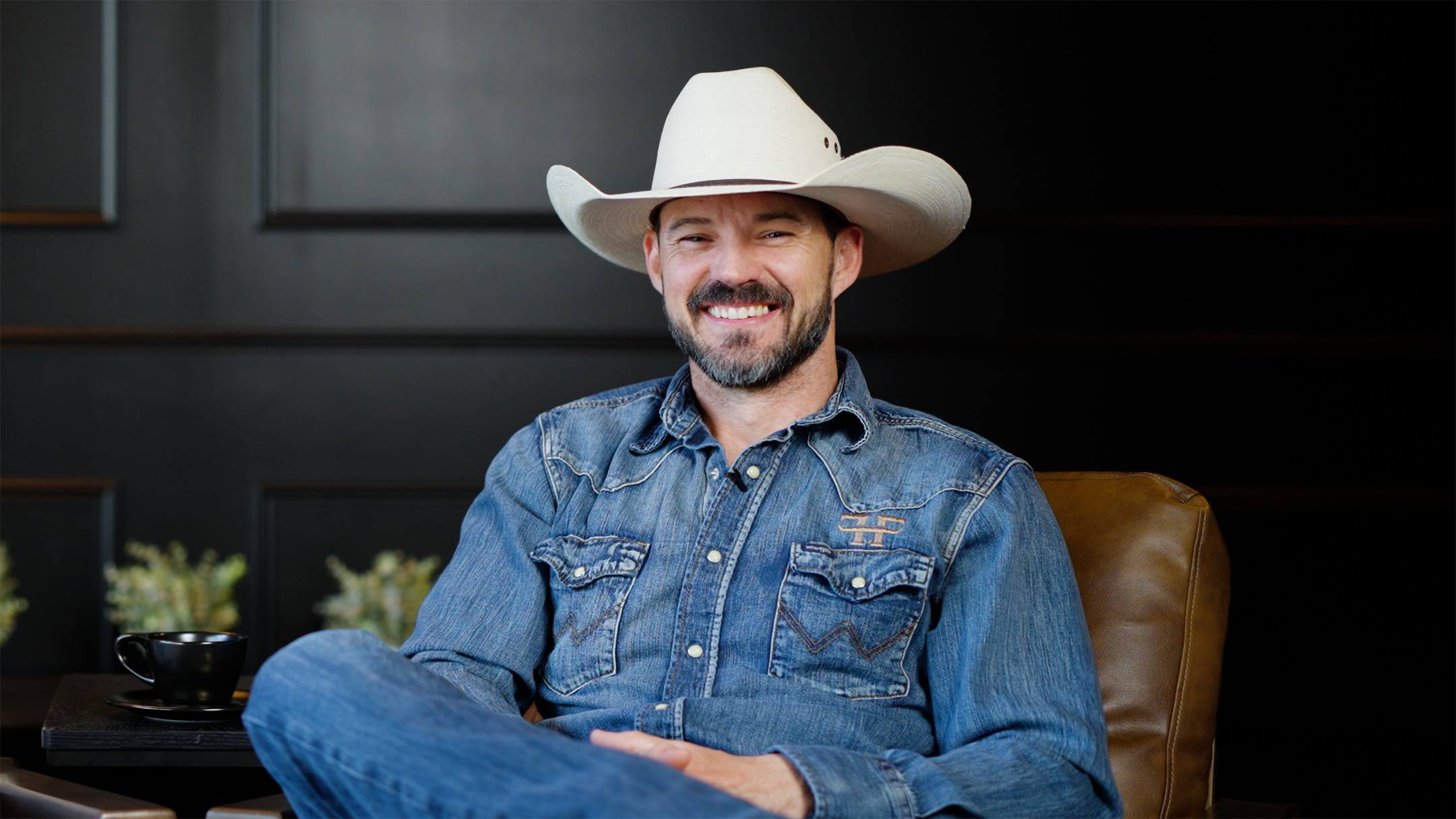 Man wearing a cowboy hat and denim shirt sitting in a chair against a dark background