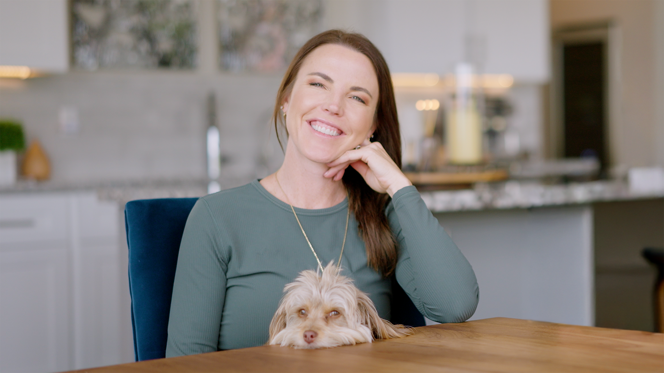 Woman sitting at a kitchen table with a small dog, smiling.