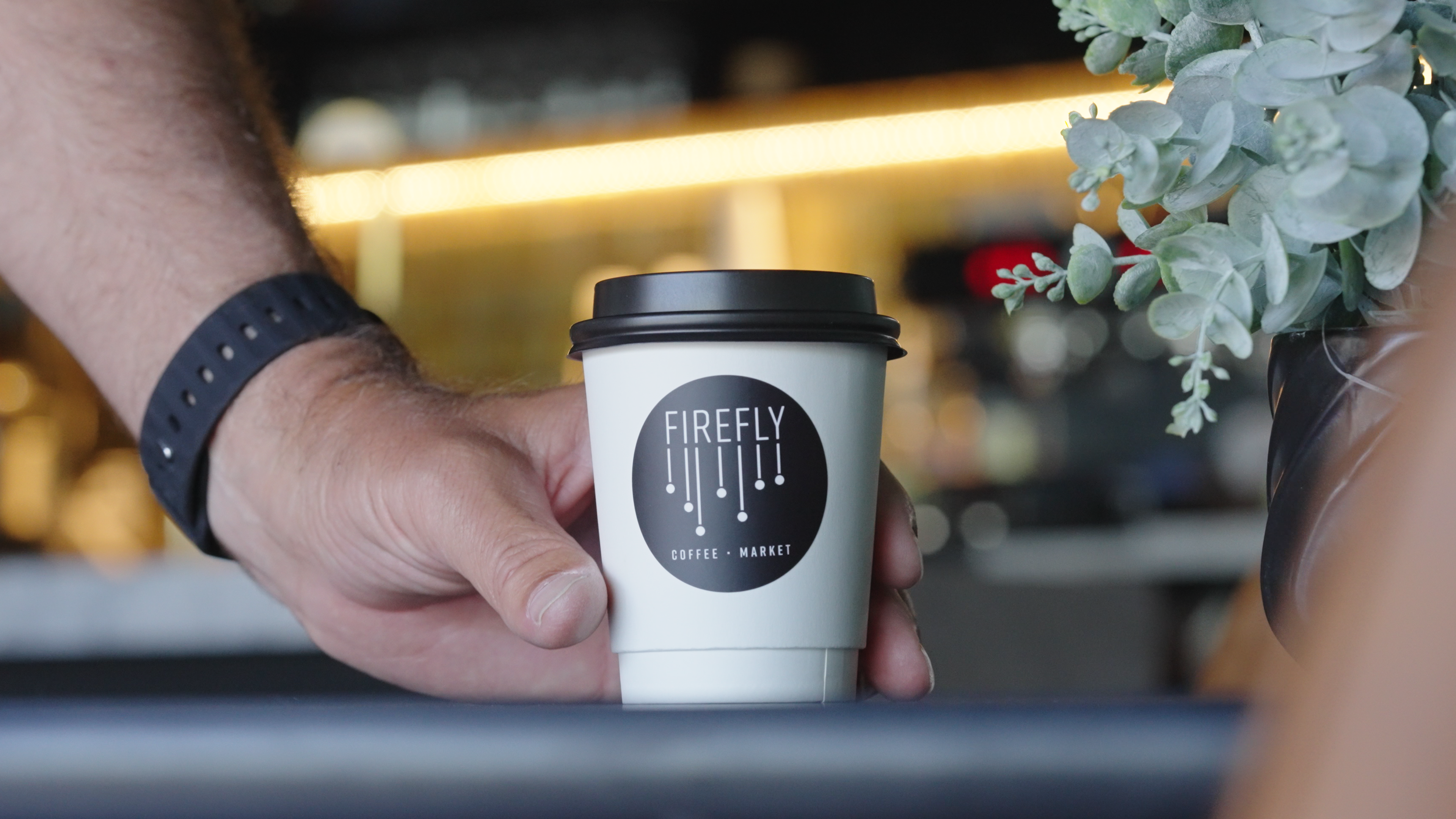 Hand holding a Firefly Coffee & Market cup with a blurred cafe background