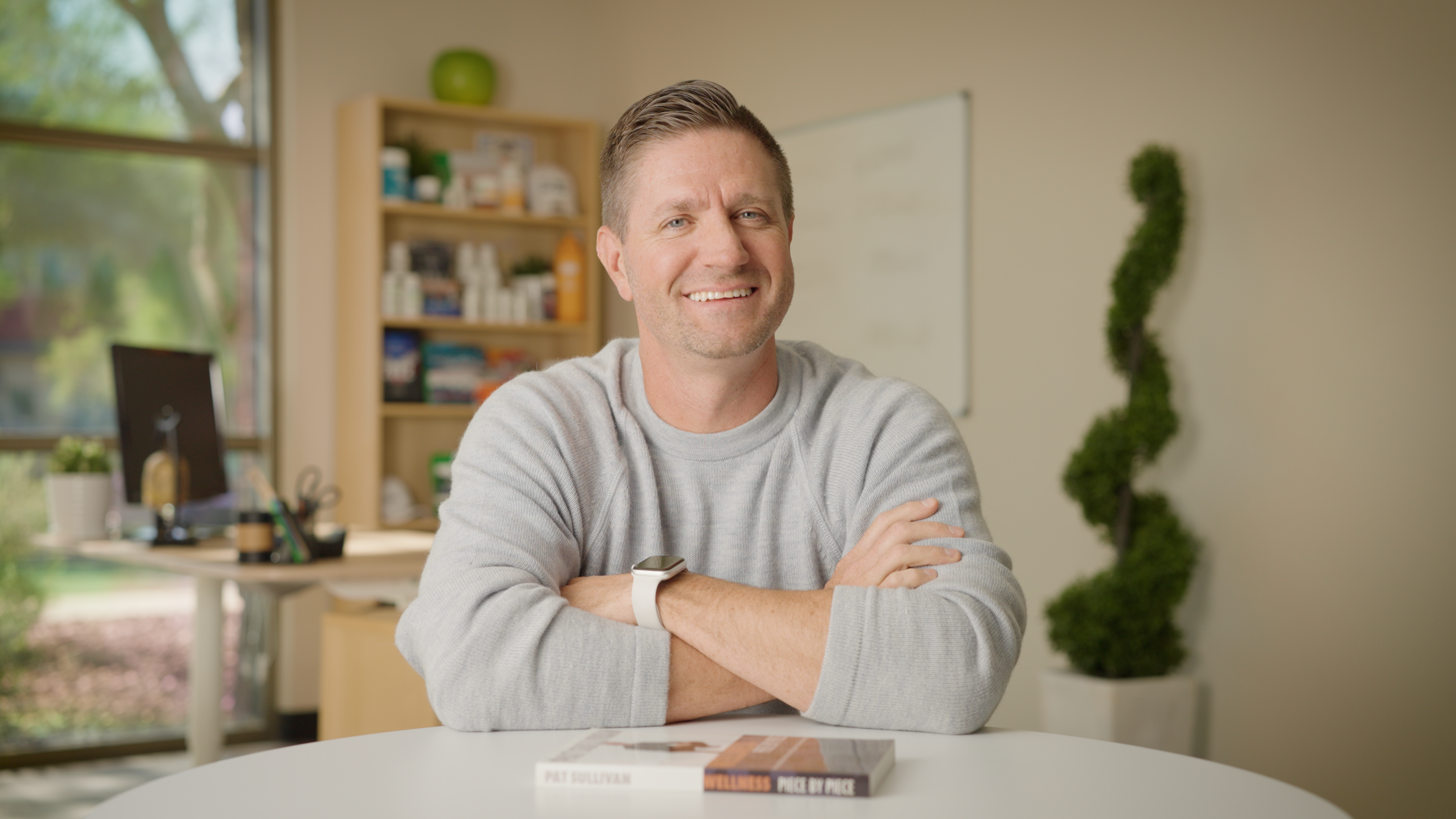 Man sitting at a table with books in a room with a window and plants.