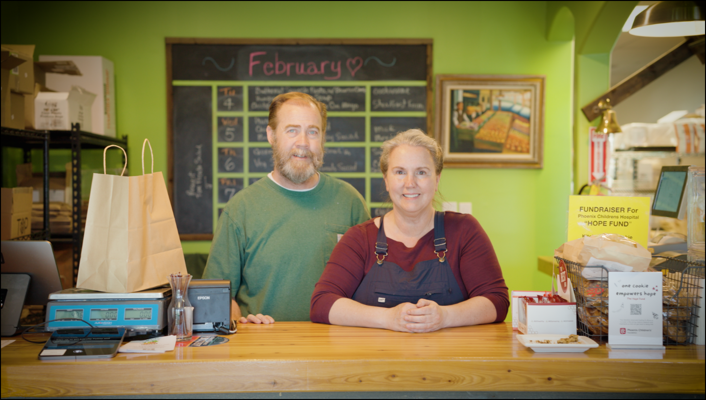 Two people standing behind a counter in a store with a green wall and calendar.