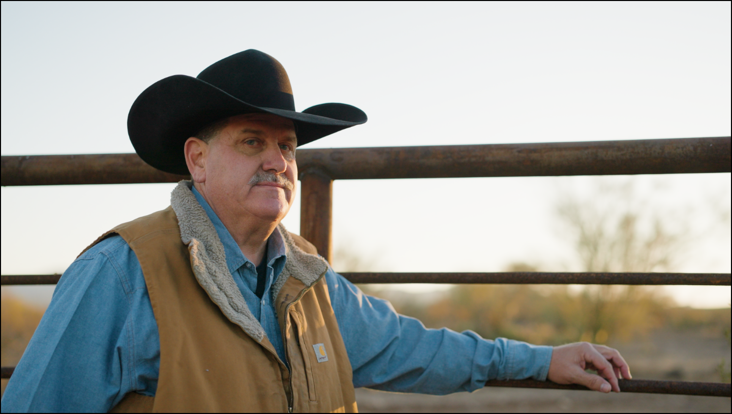 Man wearing a cowboy hat and vest standing by a fence with a blurred background
