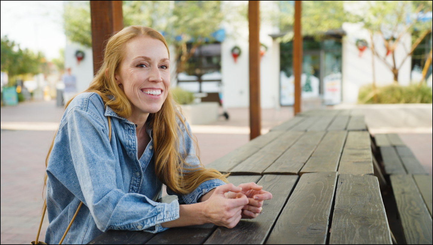 Woman sitting at a wooden table outdoors, smiling.